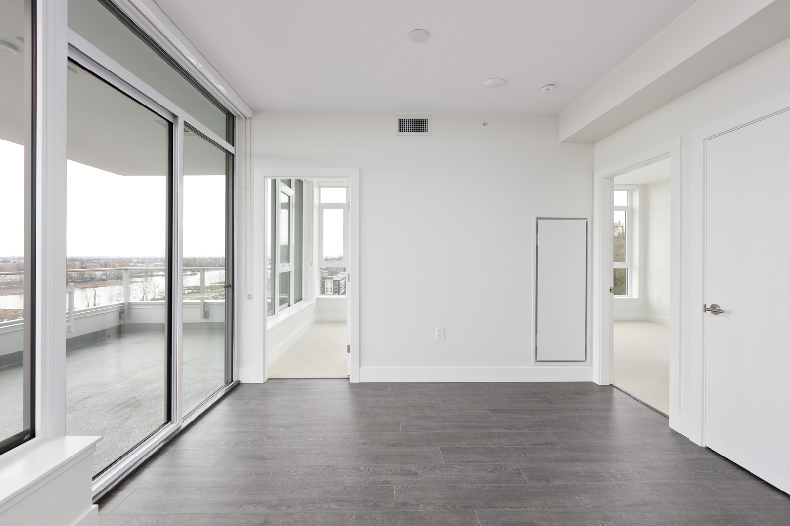 Empty room with dark wood flooring, large sliding glass doors leading to a balcony, and two doorways to other rooms. Walls are painted white, creating a bright and open space.