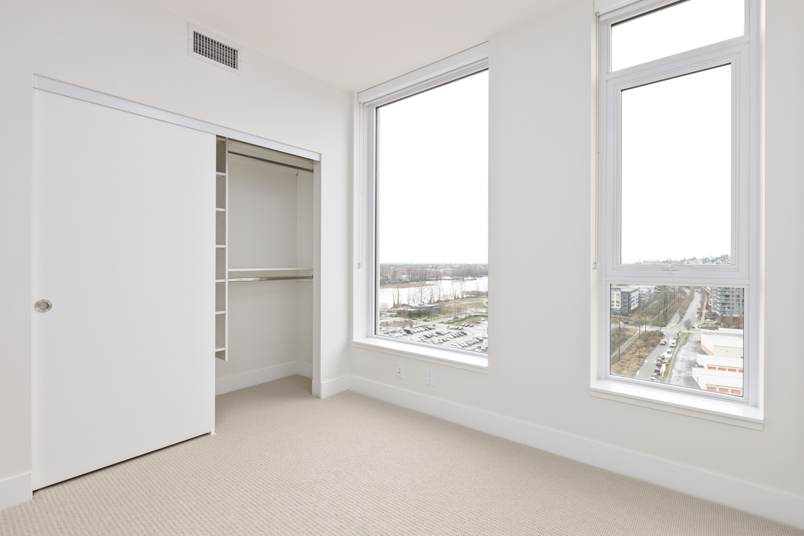 Empty room with beige carpet, an open closet with shelves and hanging space, and two large windows showing an urban and river view.