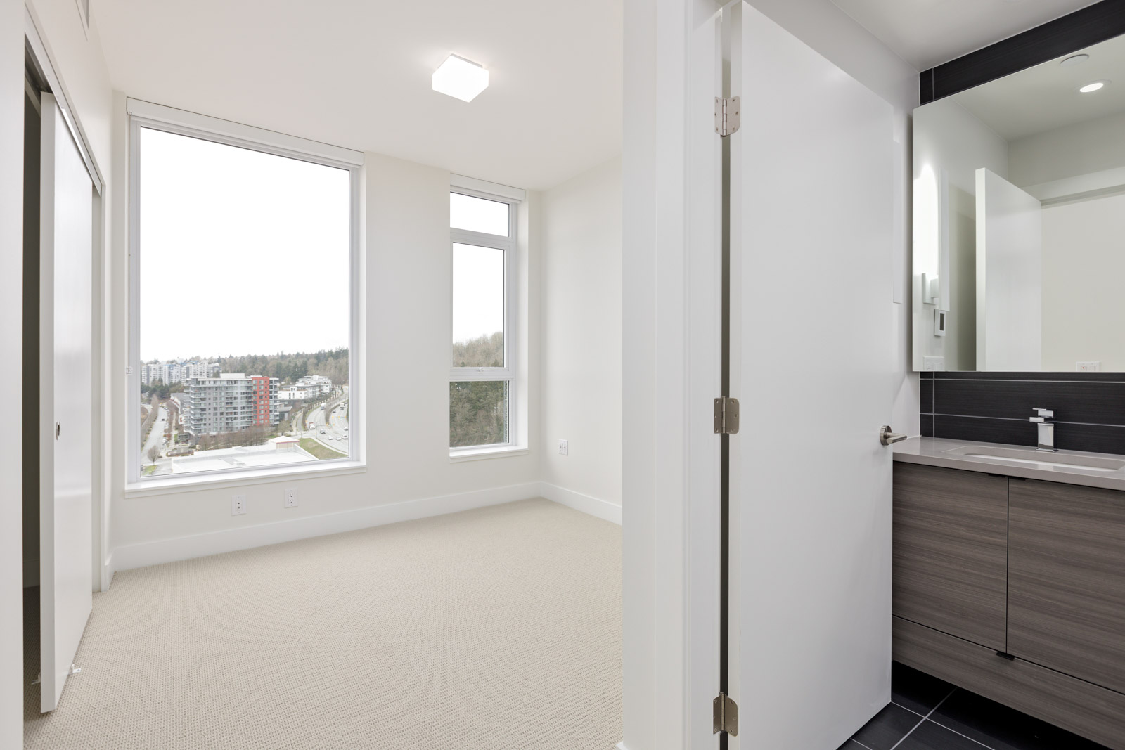 A bright, empty room with large windows next to a bathroom with a modern sink and mirror. Carpeted floor in the room and tiled floor in the bathroom.