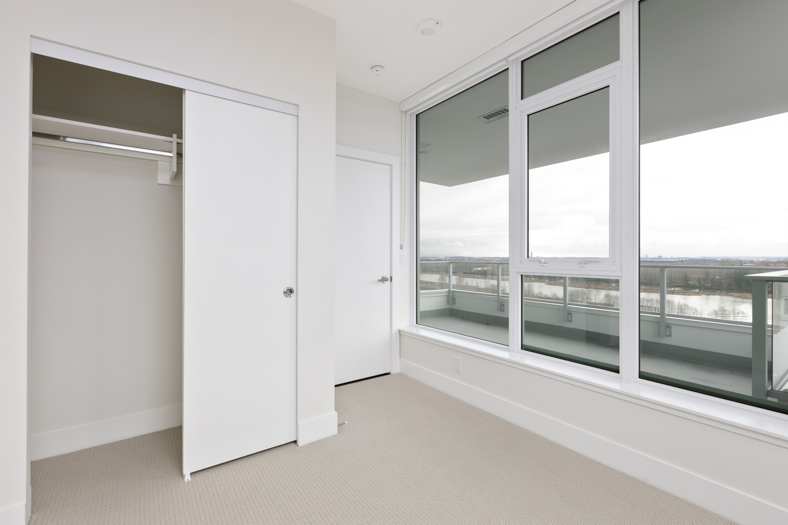 Minimalist room with beige carpet, open closet, and large windows overlooking a balcony.