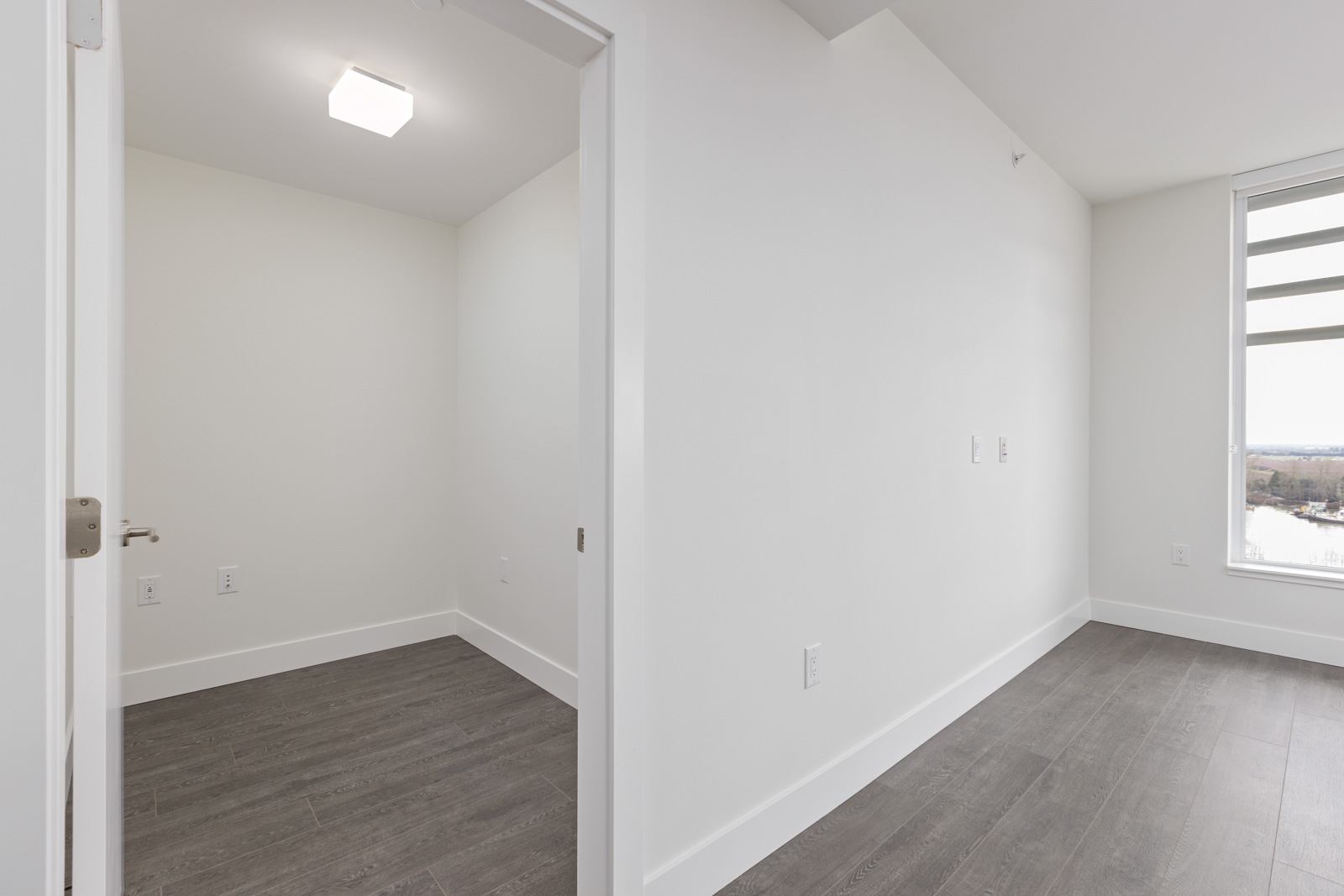 Empty room with white walls and dark wooden flooring, featuring a window with a view and an open doorway leading to a smaller adjoining space.