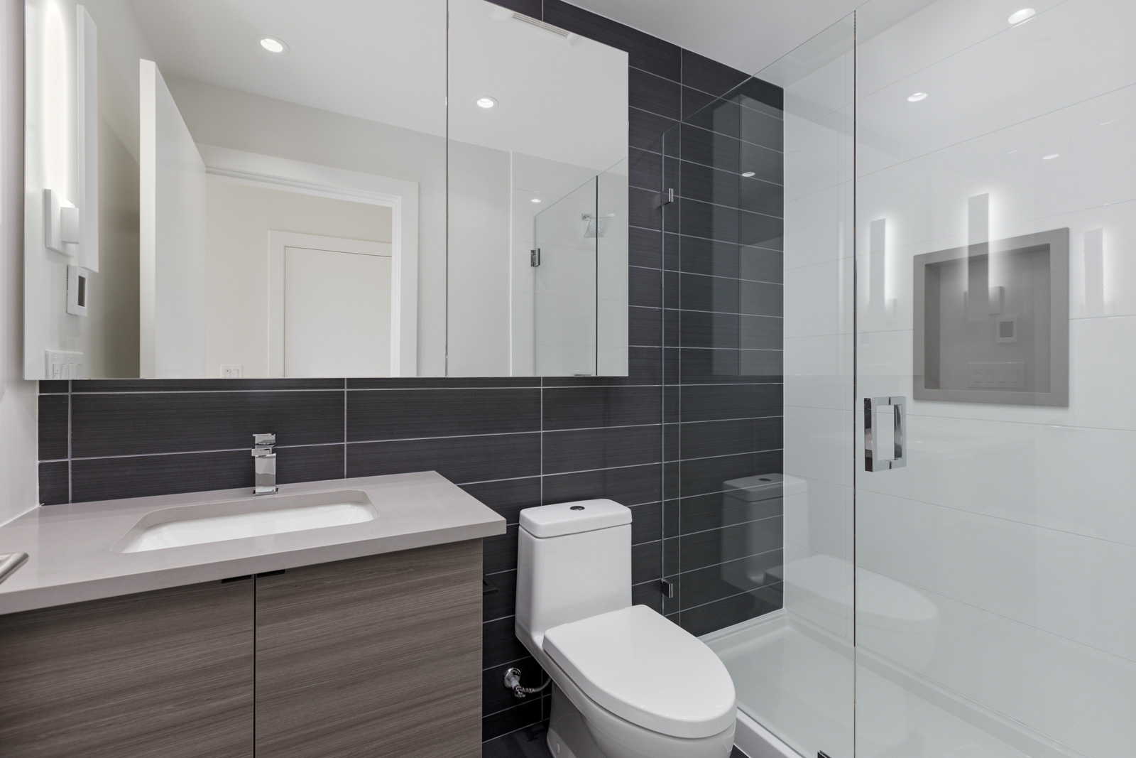 Modern bathroom with gray tile walls, a glass-enclosed shower, a toilet, and a sink with a wood-toned cabinet.