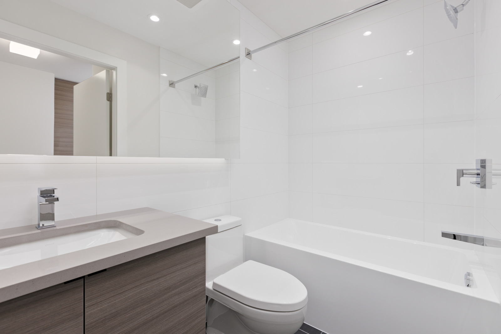 Modern bathroom with a white bathtub, toilet, and sink on a wooden vanity. Walls are tiled, and there is a large mirror above the sink. Bright lighting illuminates the space.