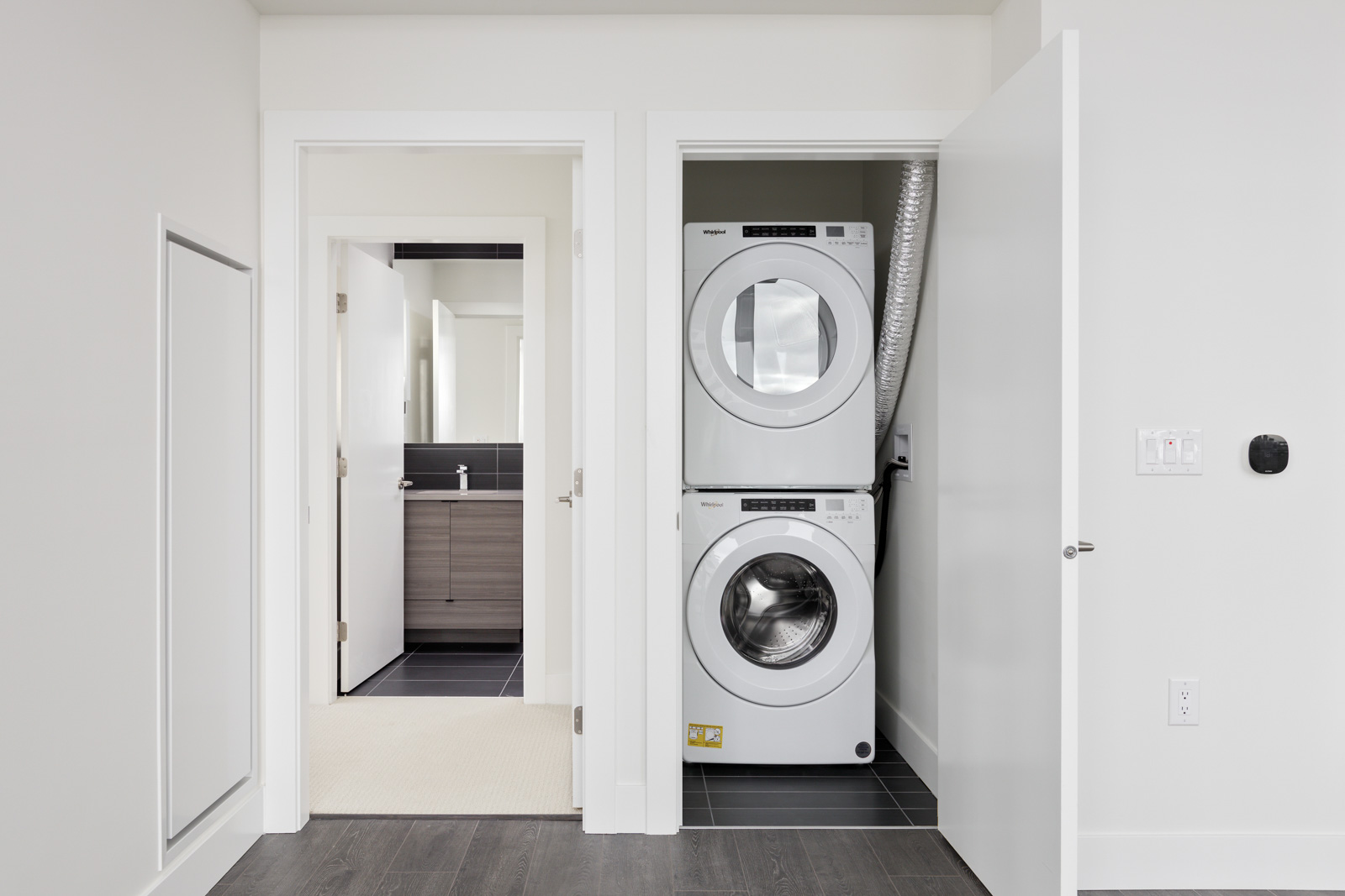 Stacked washer and dryer in a closet with white doors, adjacent to an open door leading to a bathroom with a modern vanity.