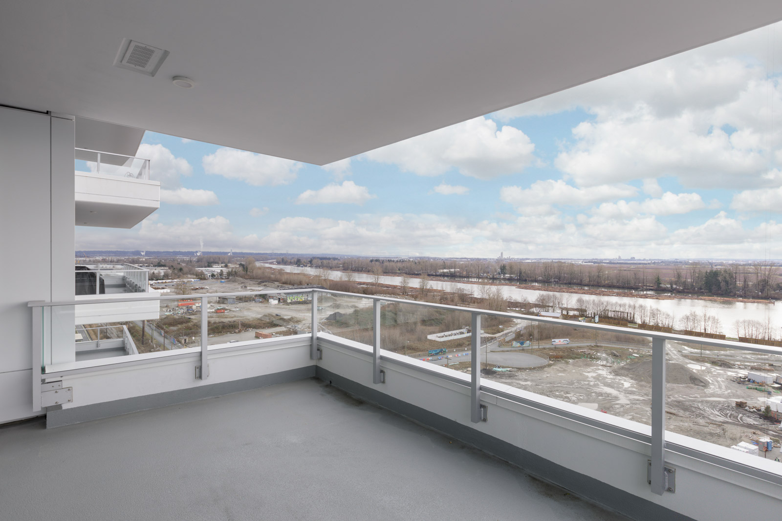 Balcony view overlooking a river, with distant trees and a partly cloudy sky. White railing and buildings are visible to the left.