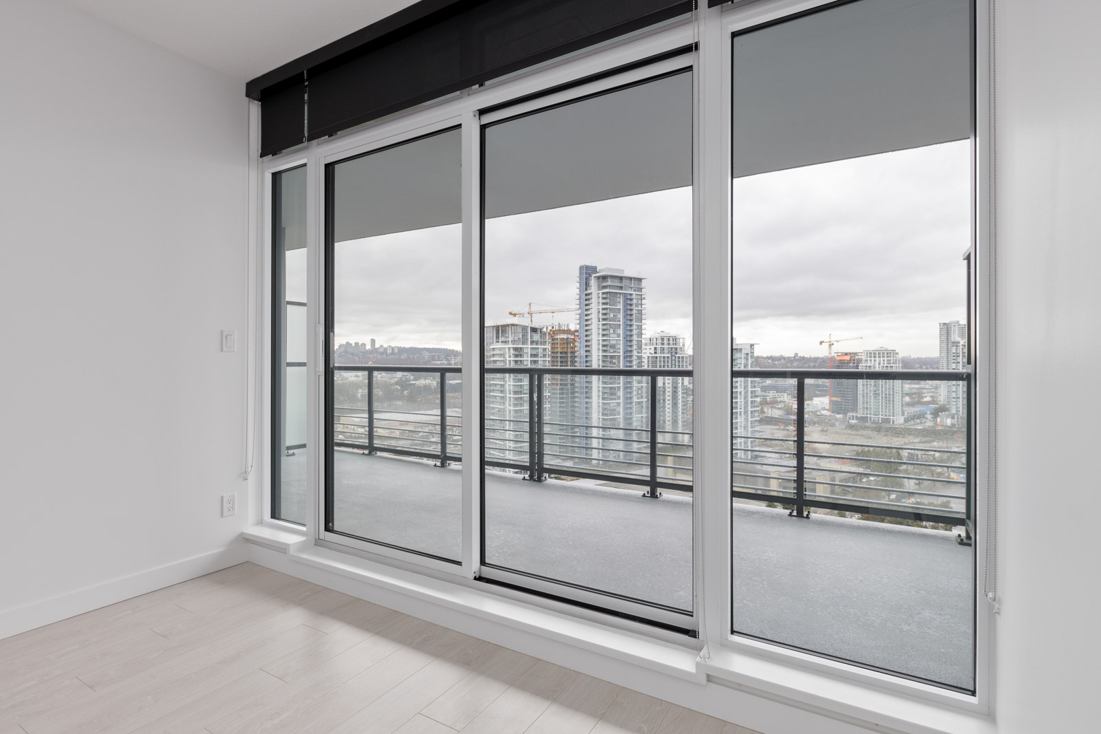 Empty modern room with light wood floors, white walls, and floor-to-ceiling windows leading to a balcony overlooking a cityscape with high-rise buildings and construction cranes.