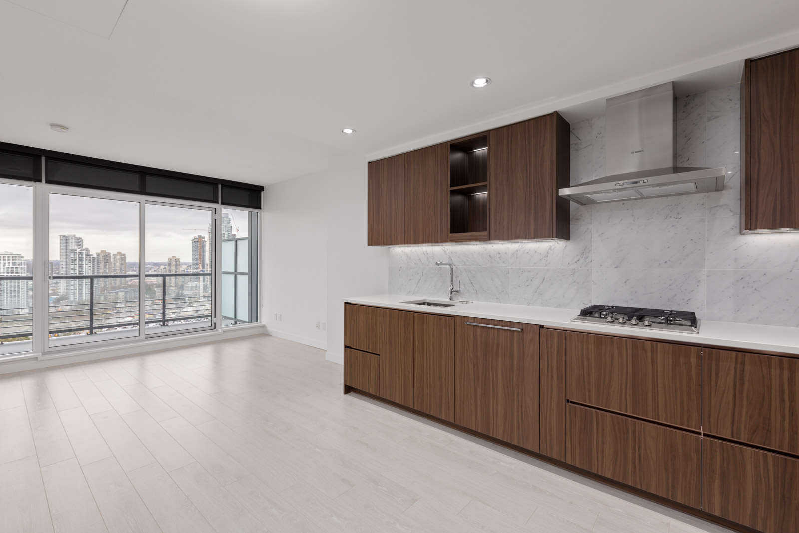 Modern kitchen with wood cabinets, marble backsplash, gas cooktop, and built-in range hood, adjacent to a bright living area with large windows and city views.
