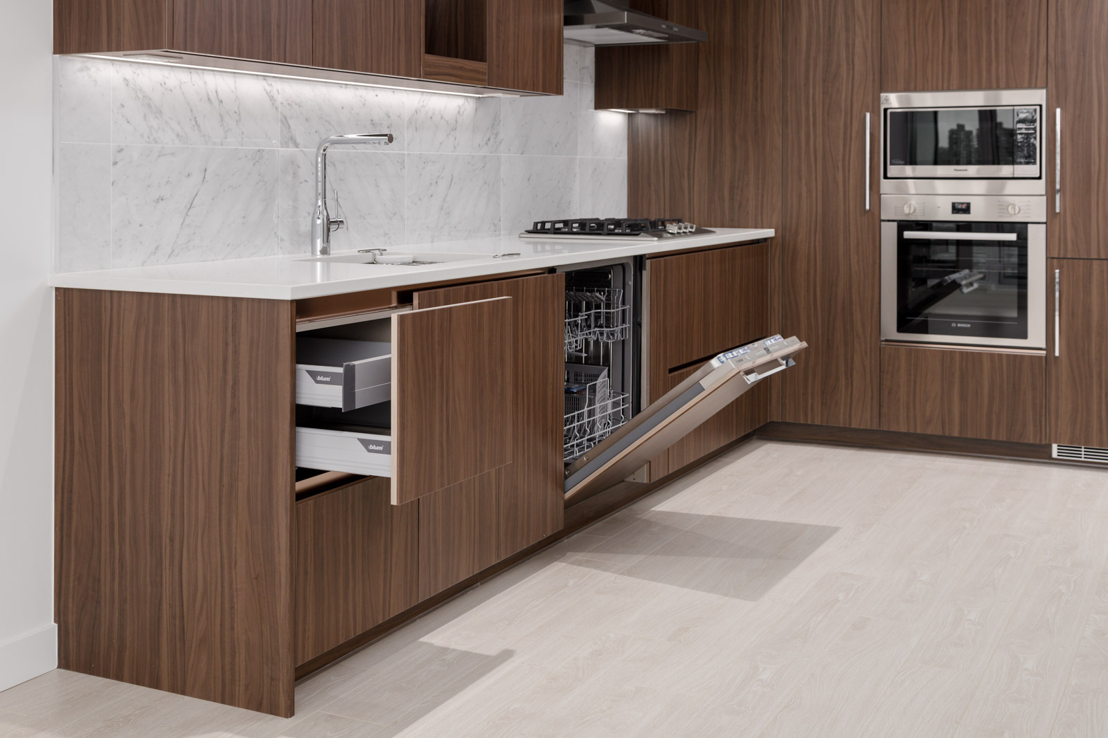 Modern kitchen with wood cabinets, open drawers, dishwasher door down, built-in oven and microwave, marble backsplash, and light wood flooring.