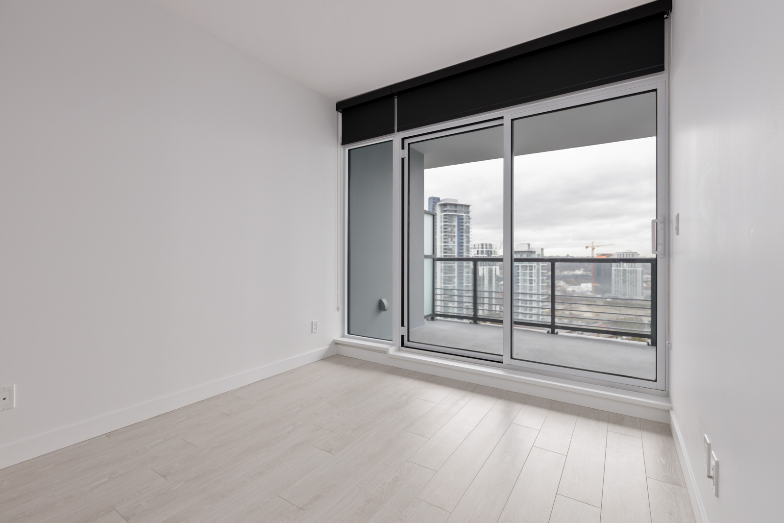 Empty modern room with light wood flooring, white walls, and large sliding glass doors opening to a balcony with a cityscape view.