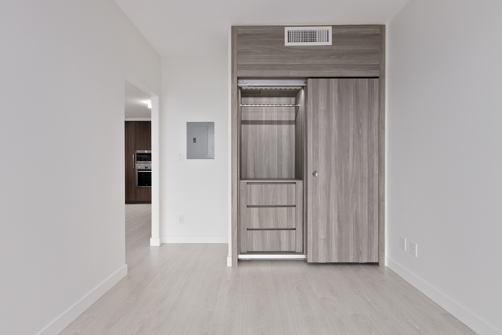 Empty room with light wood flooring, a built-in closet with sliding doors, and a view into another room with kitchen appliances visible in the background.