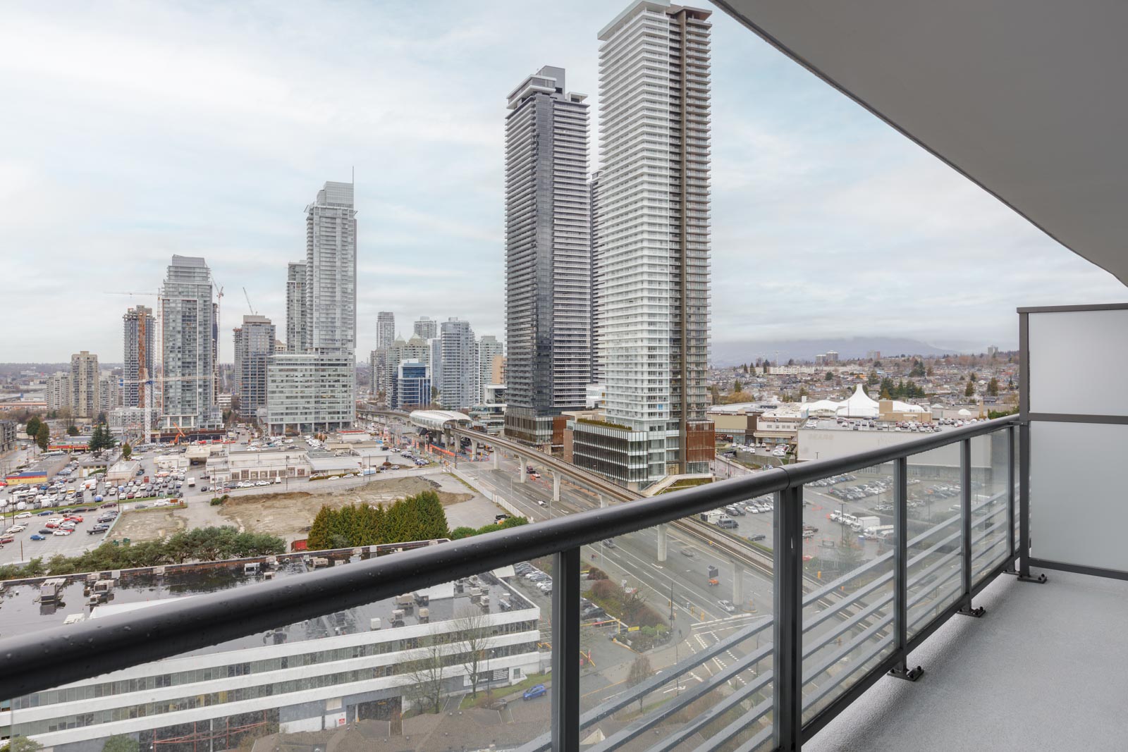 View from a high-rise balcony overlooking a cityscape with tall buildings, a construction site, roads, and a cloudy sky in the background.