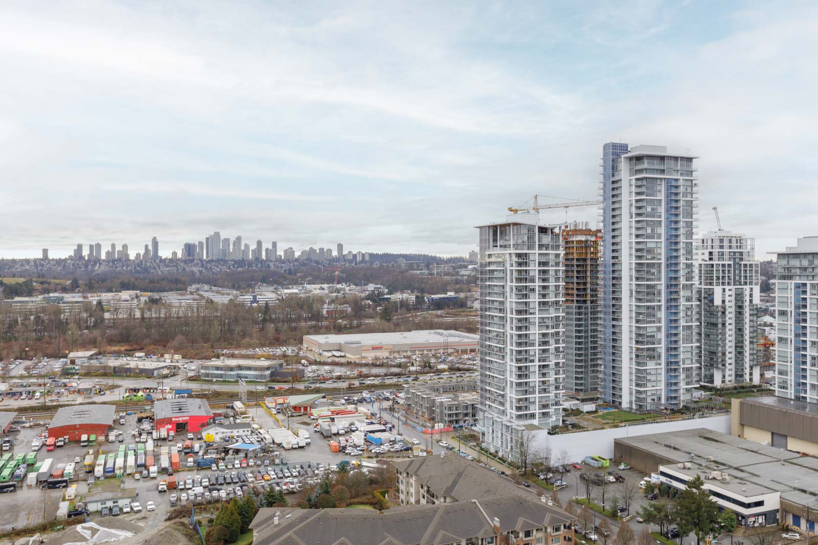 Aerial view of a cityscape with high-rise buildings, construction cranes, industrial lots, and a distant skyline under a cloudy sky.