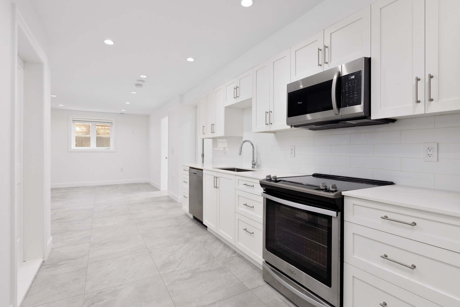 Modern kitchen with white cabinets, stainless steel appliances, including a microwave and oven, tiled backsplash, and gray tiled floor, leading to a bright open space in the background.