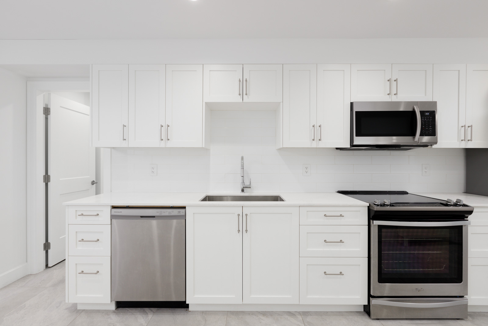 A modern kitchen with white cabinets, stainless steel appliances including a dishwasher, microwave, and oven, a double sink, and a white tile backsplash. There is a door to the left.
