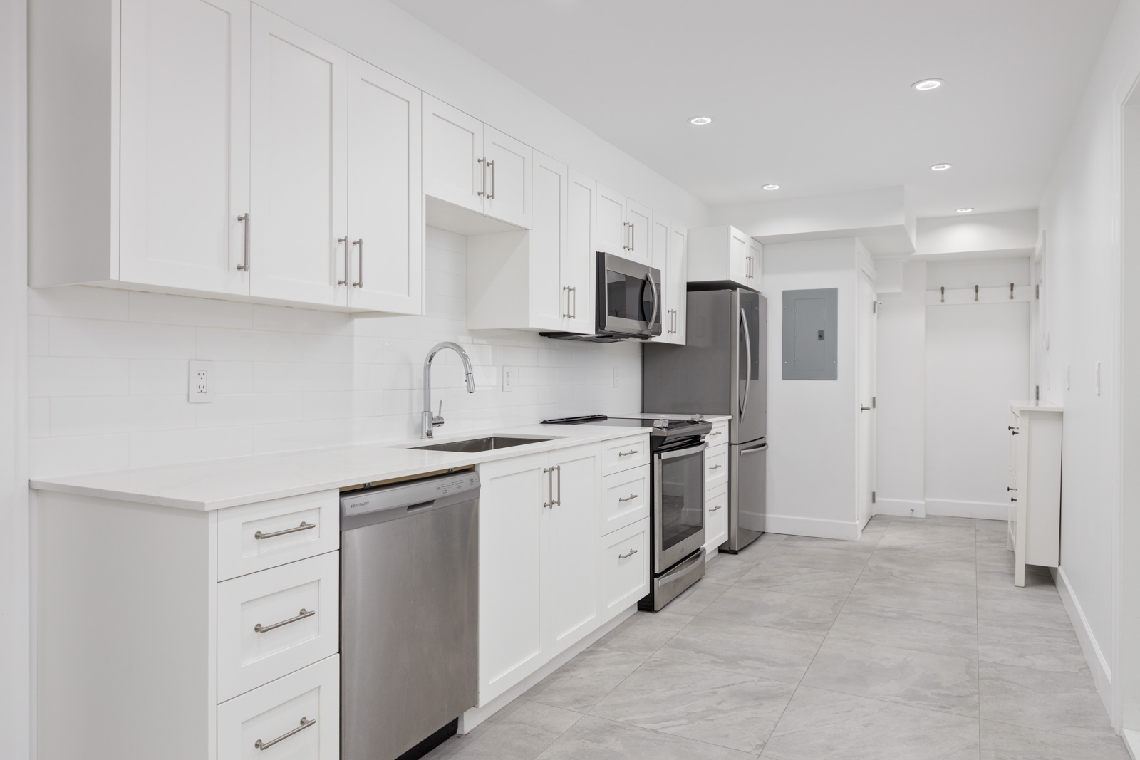 A modern kitchen with white cabinets, stainless steel appliances, a tiled backsplash, and gray tile flooring. The kitchen includes a sink, dishwasher, oven, microwave, and refrigerator.