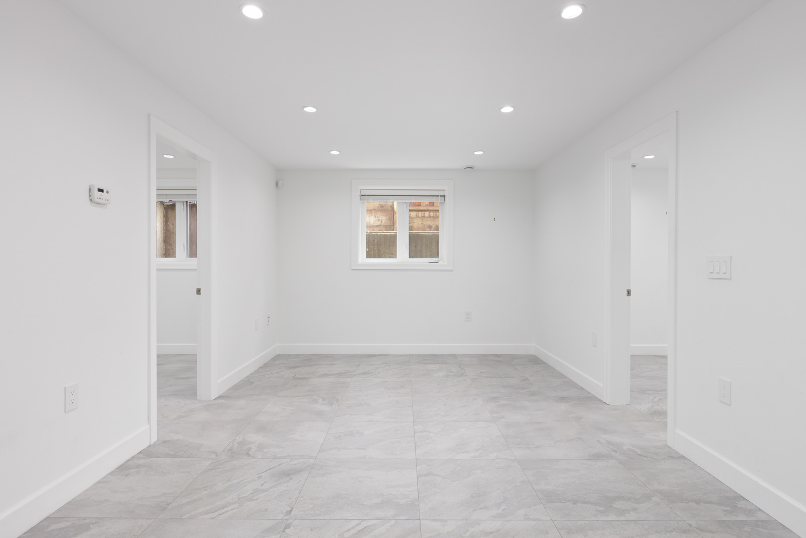 A clean, empty room with white walls, a window in the back, and tiled flooring. The room has three doorways, recessed lighting, and light switches on the wall.