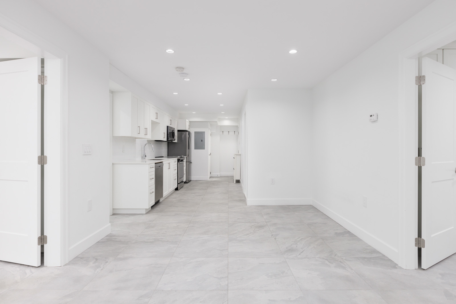 A modern, unfurnished apartment kitchen with white cabinets, stainless steel appliances, and light gray tiled flooring. The space is well-lit with recessed lighting and has a clean, minimalist design.