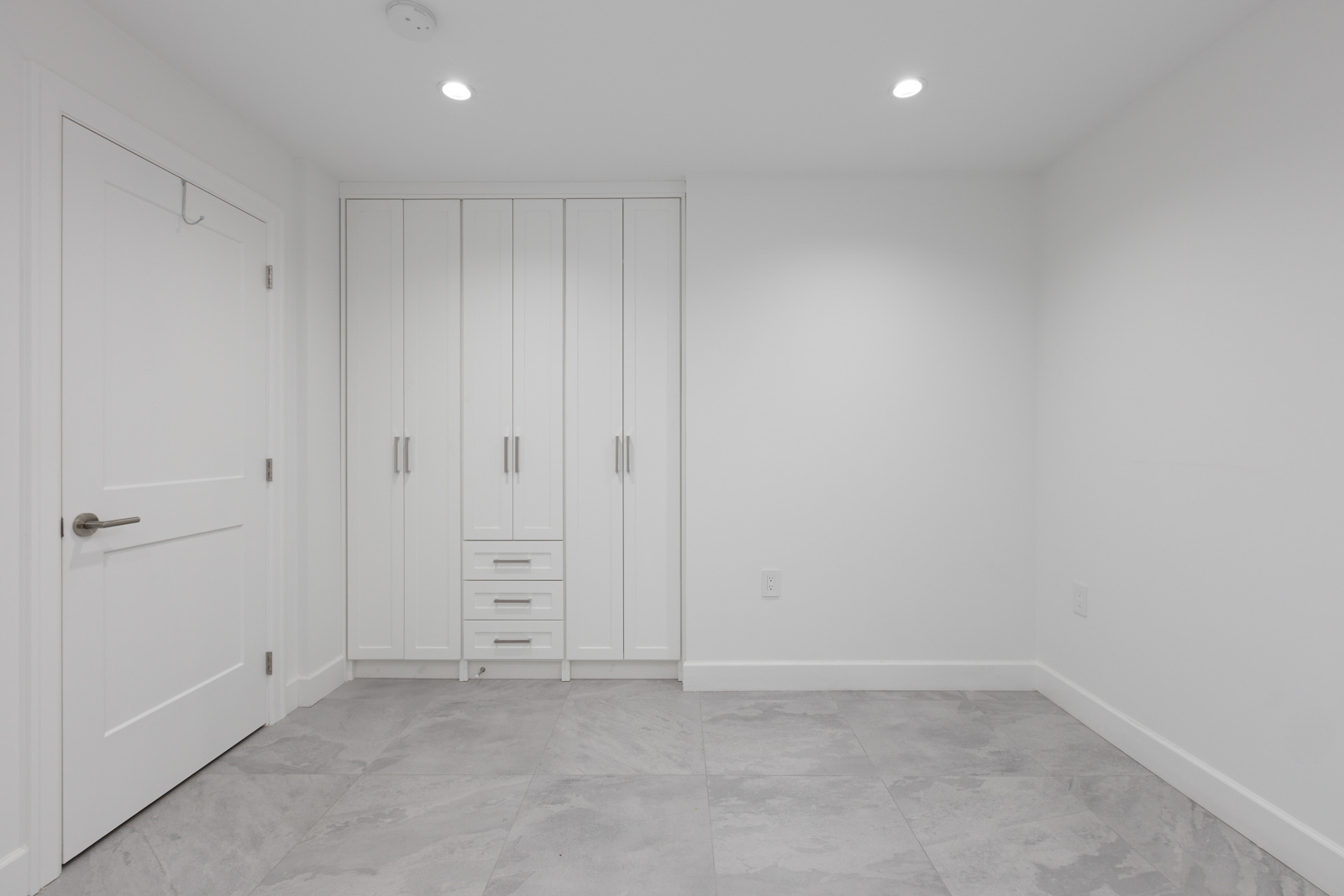 A clean, empty room with tiled flooring, a door on the left, and a built-in wardrobe with cabinets against the far wall.