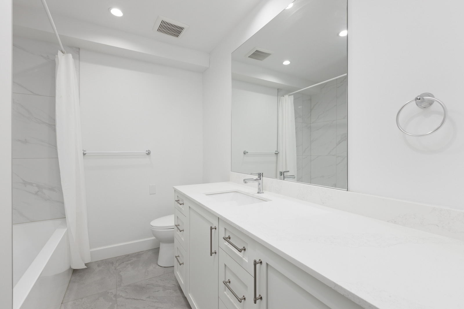 Modern white bathroom with a large mirror, single sink with a faucet, white countertop, drawers, a towel ring, and a bathtub with a shower curtain. Walls and floor have light-colored tiles.