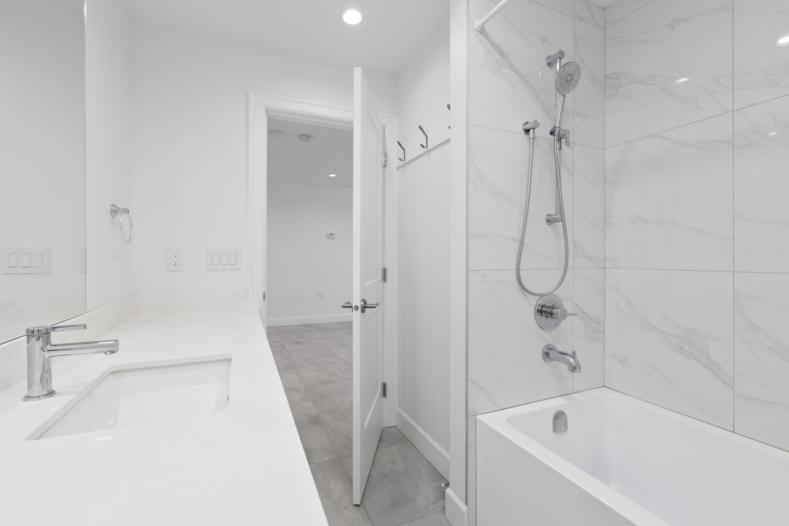A modern bathroom with a white countertop, sink, wall-mounted faucet, tiled bathtub, and showerhead. The door is open, leading to a room with tiled flooring.