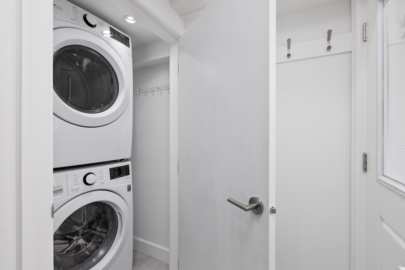 A small laundry room with a stacked washer and dryer set, a partially open white door, and wall hooks behind the door.