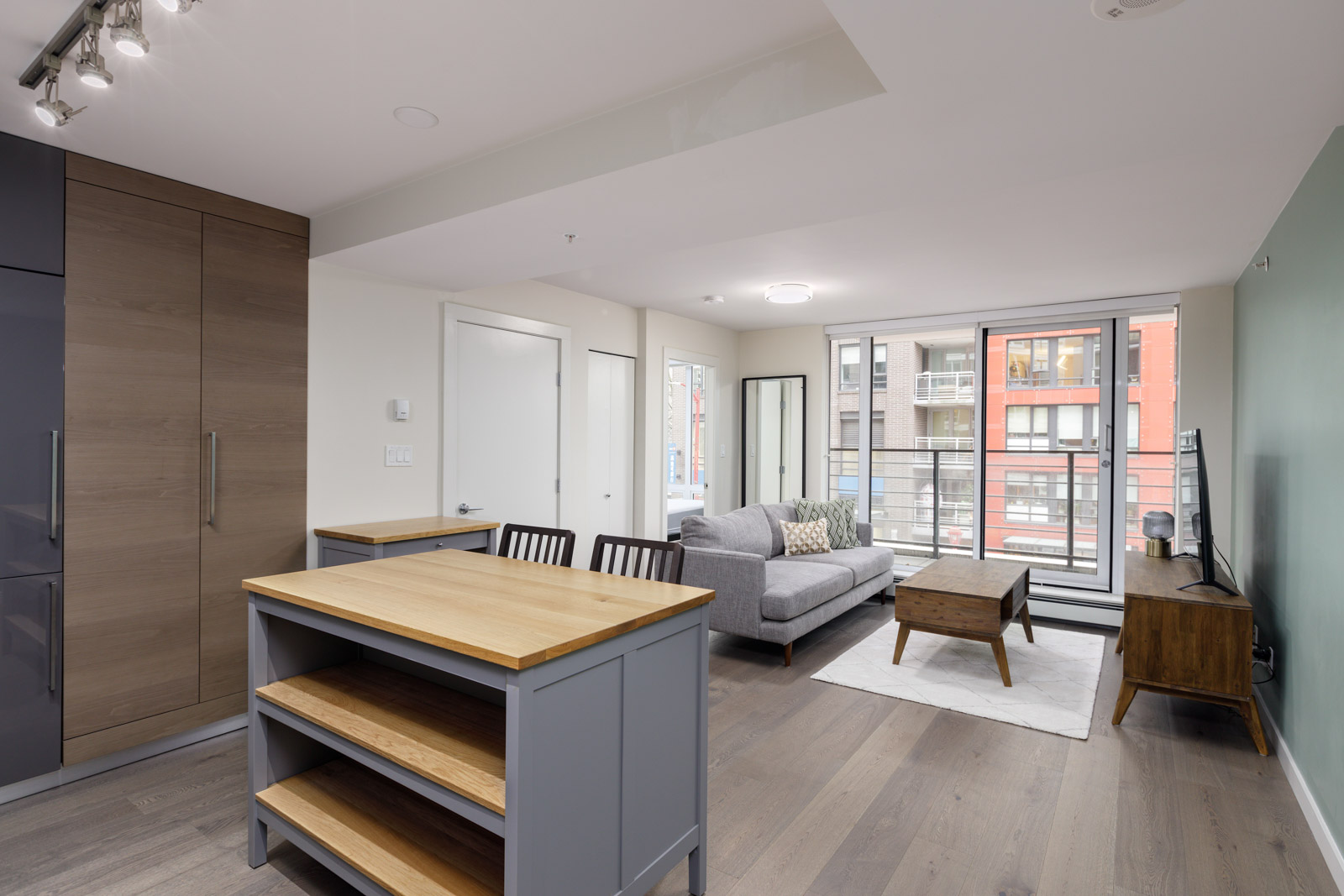 Modern open-concept living room and kitchen with light wooden flooring, gray sofa, wooden table, and green accent wall. Large windows reveal city buildings outside.