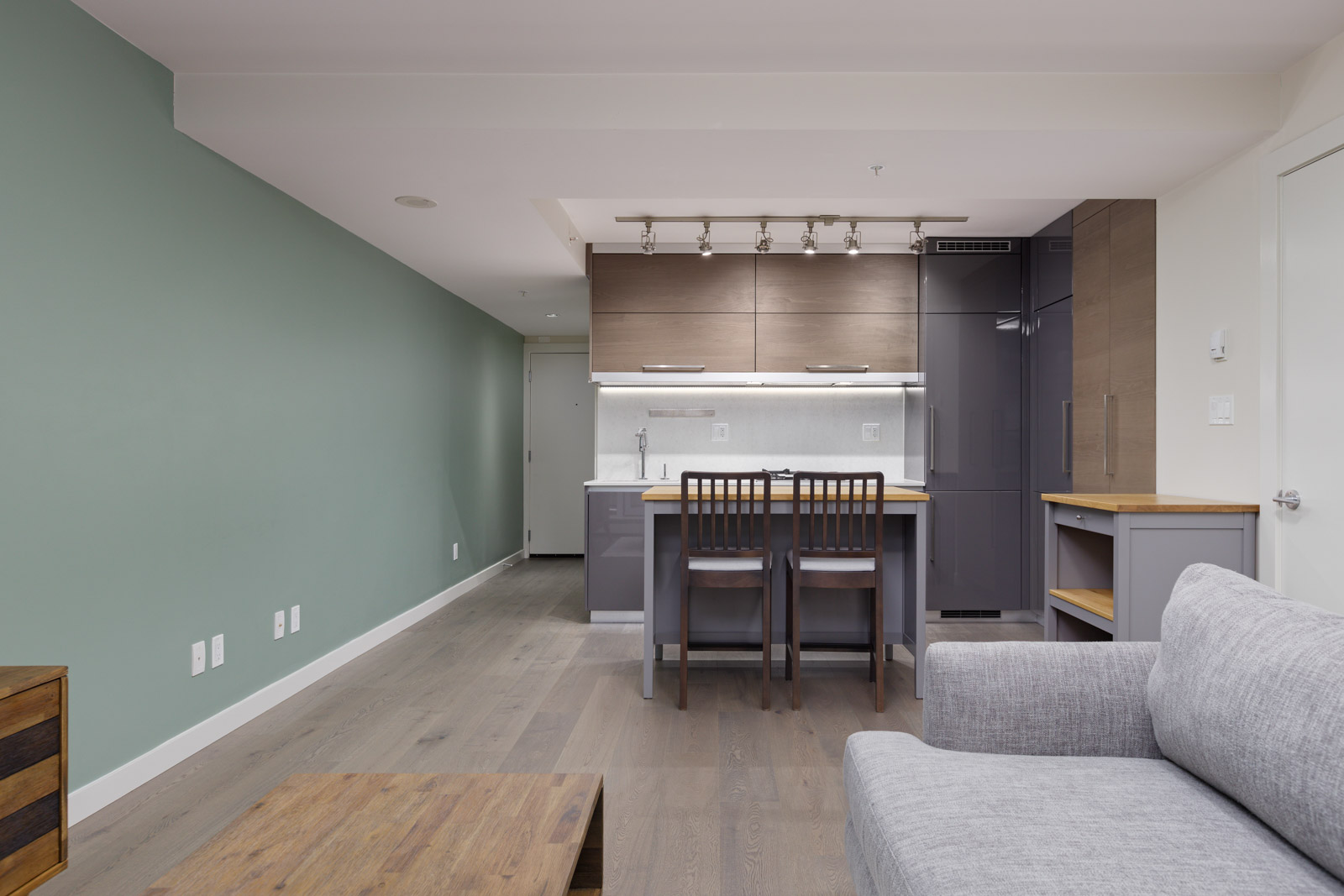 Modern open-plan apartment with a minimalist kitchen and living area. Features light wood flooring, green accent wall, gray sofa, bar counter with two stools, and overhead cabinets.