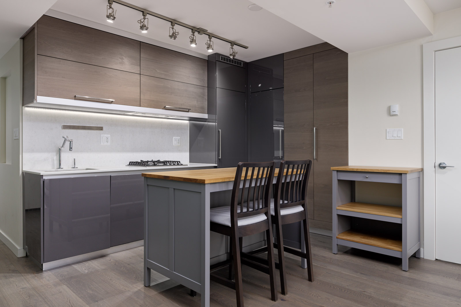 Modern kitchen with dark wood cabinetry, an island with two chairs, and stainless steel appliances. A wooden shelf stands on the right.