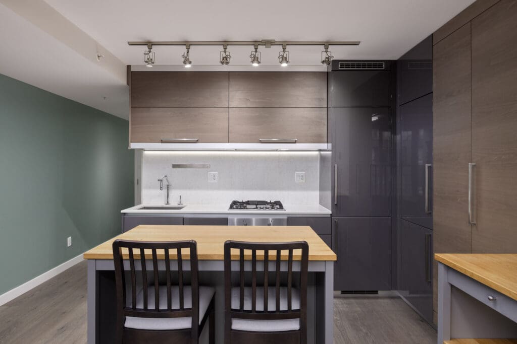 Modern kitchen with wooden cabinets, overhead lighting, and an island with two chairs. Features include a built-in stovetop and stainless steel faucet against a green accent wall.