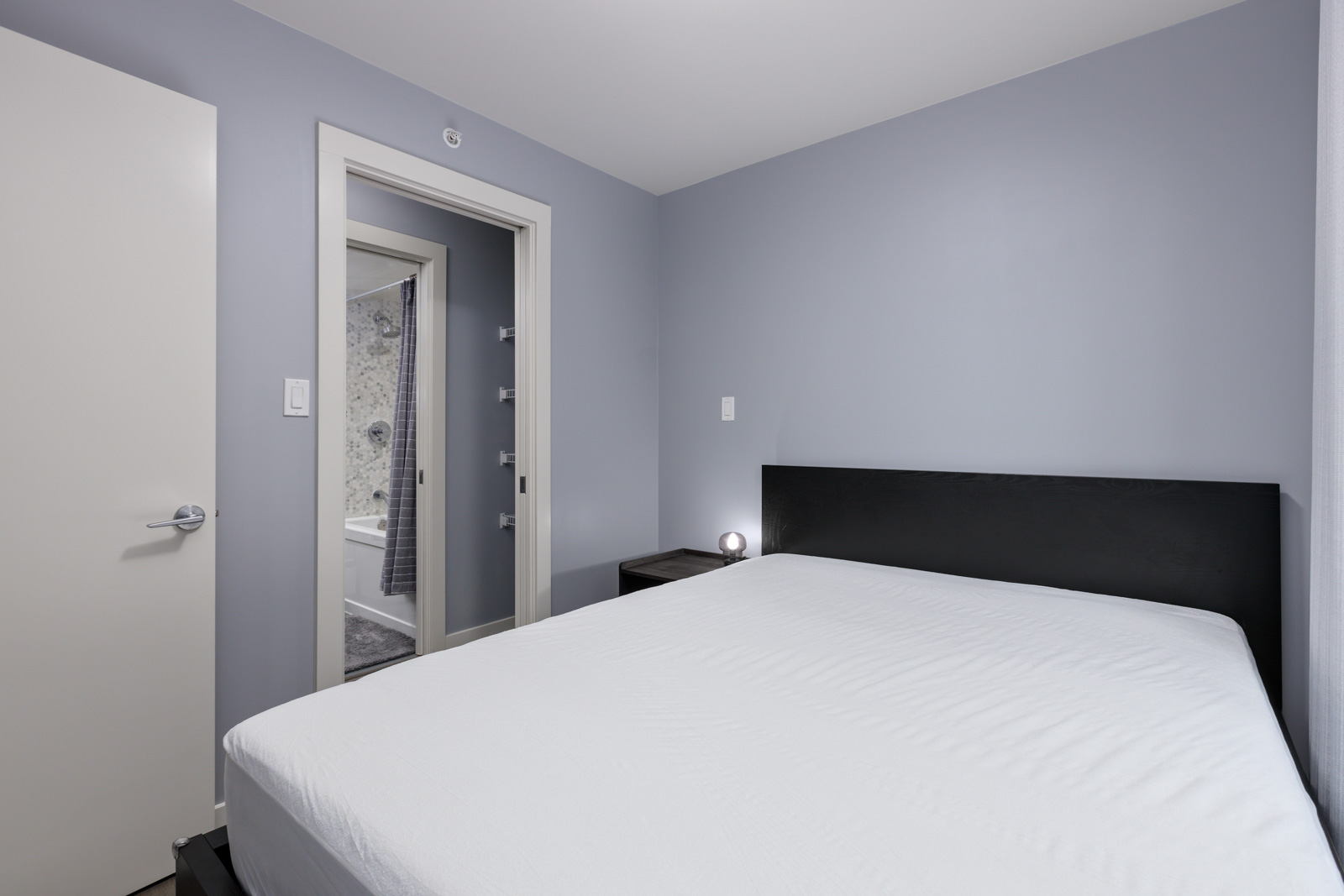 A minimalist bedroom with a bed, gray walls, and an open door showing a glimpse of a bathroom.