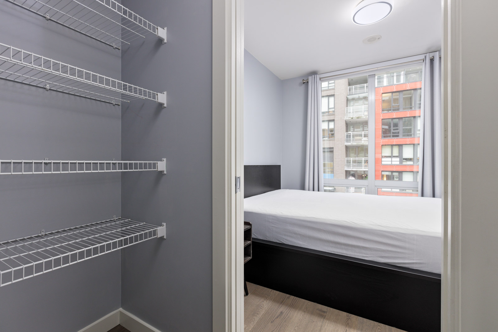 A small bedroom with a bed and gray walls, adjacent to an empty closet with wire shelving. A window shows part of a building exterior.