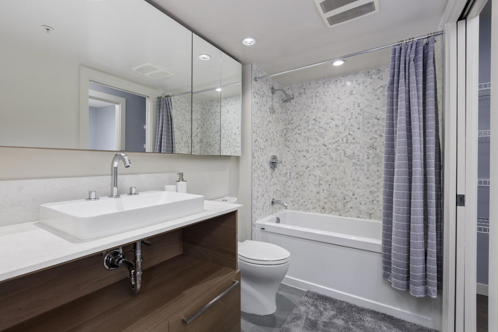 Modern bathroom with a large mirror, double sink, toilet, bathtub, and gray patterned shower curtain. Marble tiled wall and gray floor mat complete the design.