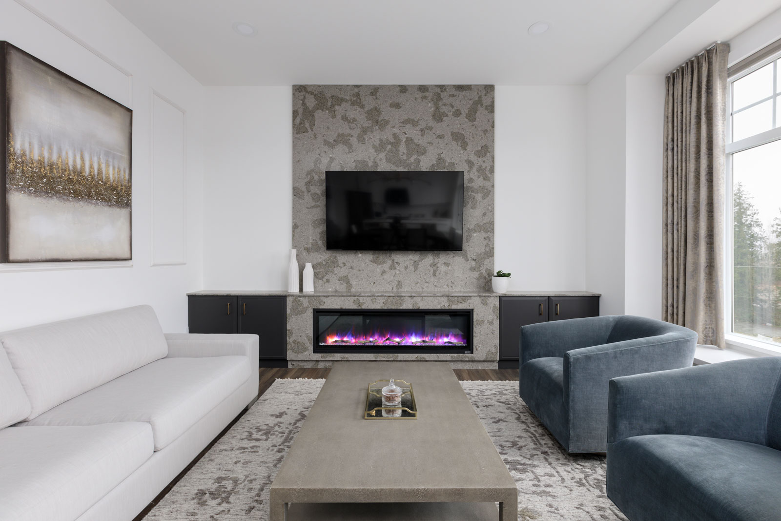 Modern living room with a white sofa, two blue chairs, a large coffee table, wall-mounted TV, electric fireplace, abstract art, and a large window with curtains.
