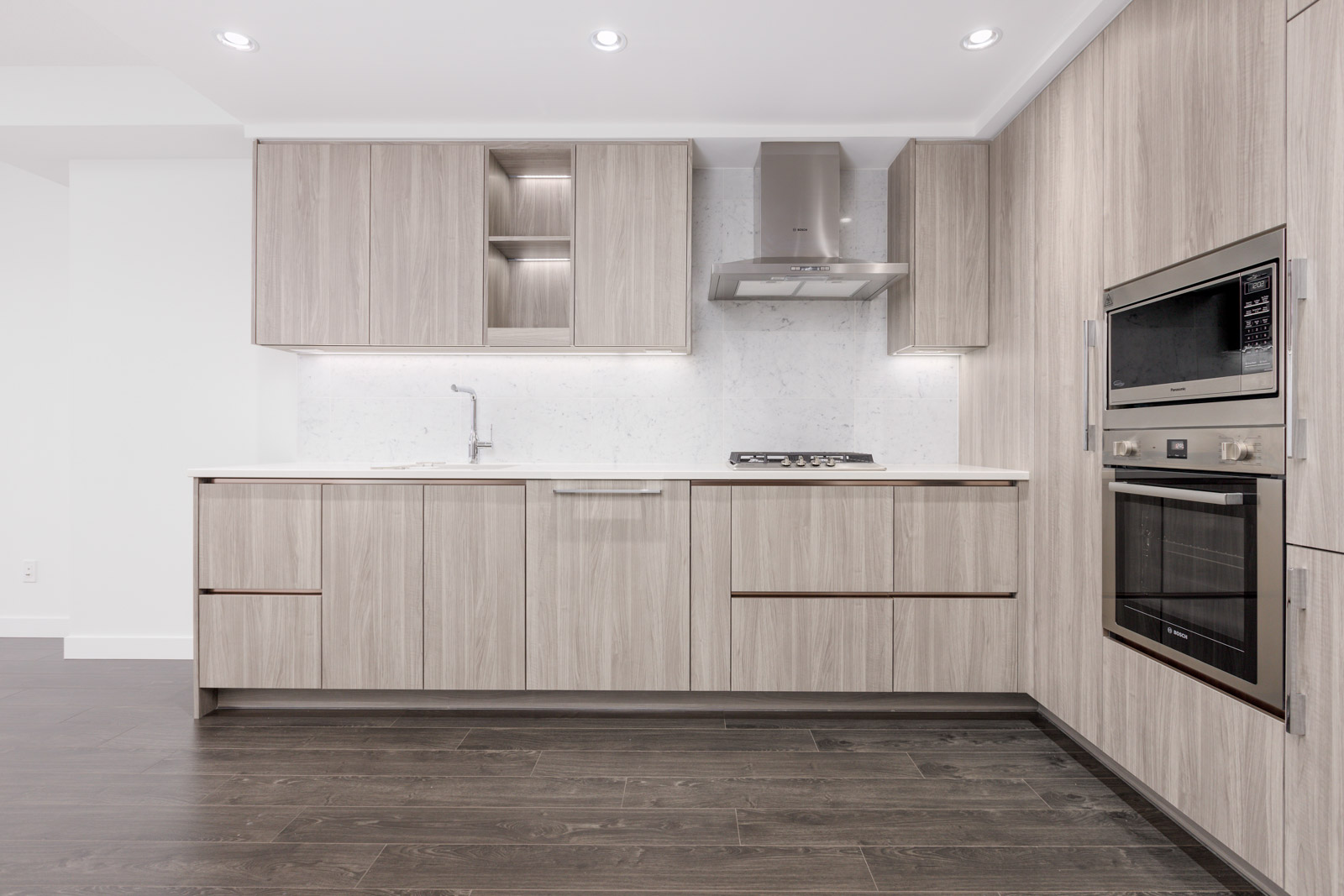 Modern kitchen with light wood cabinetry, stainless steel oven, and stovetop. Dark wood flooring contrasts with white walls and counters.