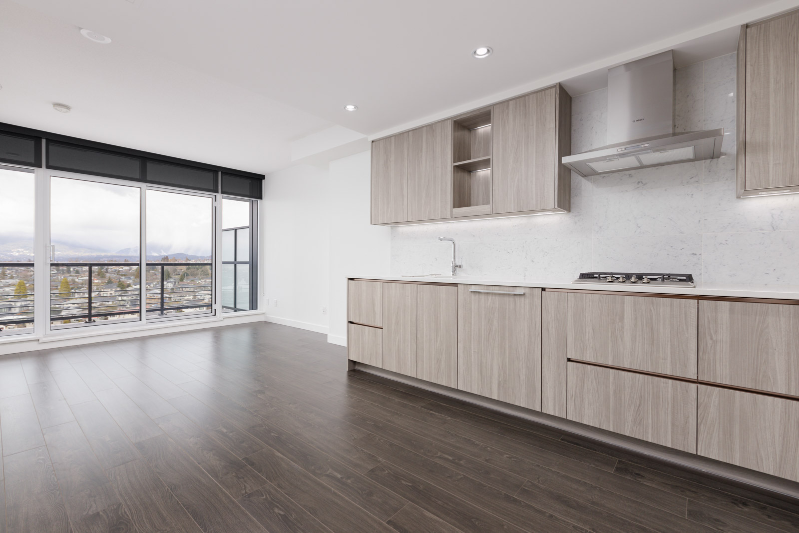 Modern kitchen with light wood cabinets, marble backsplash, and built-in stove. Hardwood floors and large windows offer natural light and a view of the cityscape.