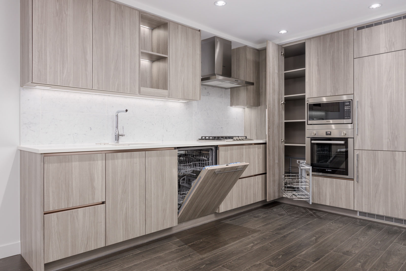 Modern kitchen with light wood cabinetry, built-in oven, dishwasher, and open cabinets. White countertops and backsplash, stainless steel appliances, and dark wood flooring.