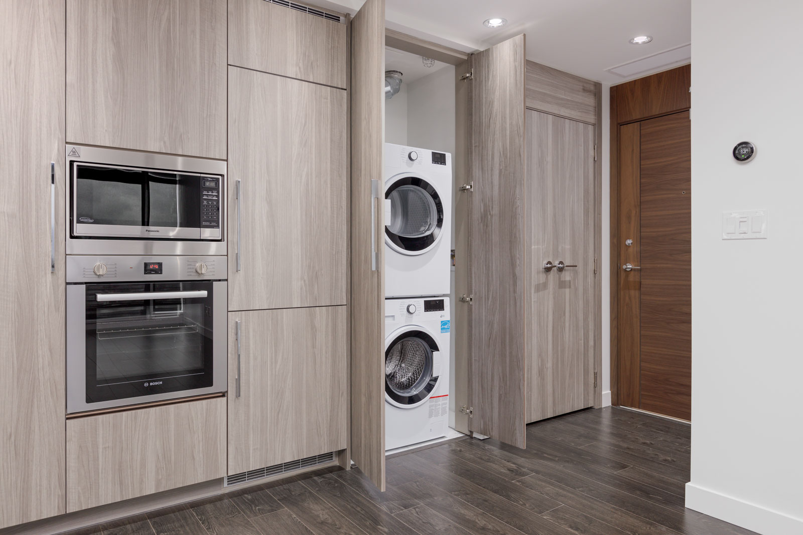 Built-in kitchen with wood cabinets, wall oven, microwave, and an open closet revealing a stacked washer and dryer. Dark wooden floor and recessed lighting visible.