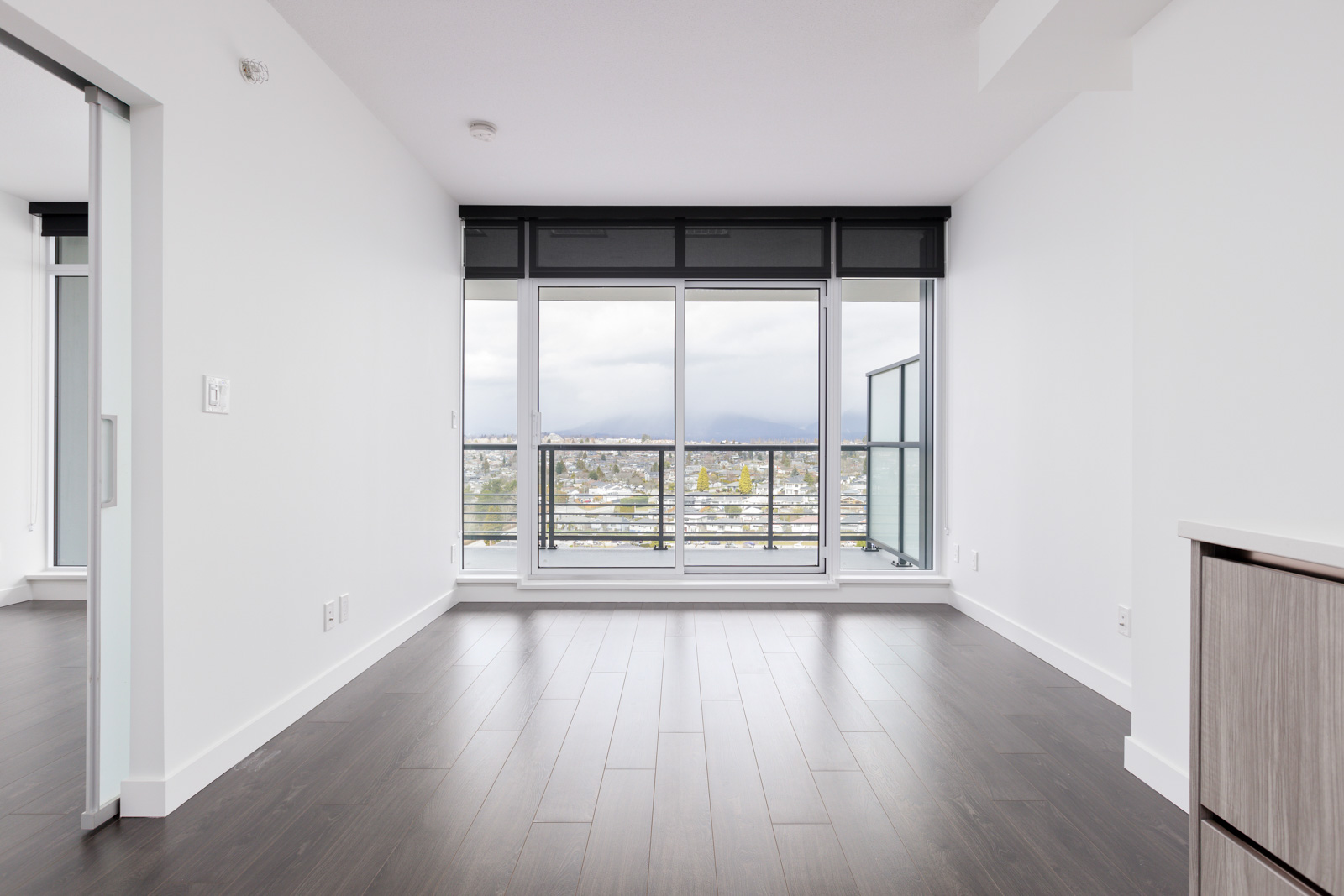 Modern empty room with dark wood flooring, large windows, and city view. White walls and sliding door to the left.