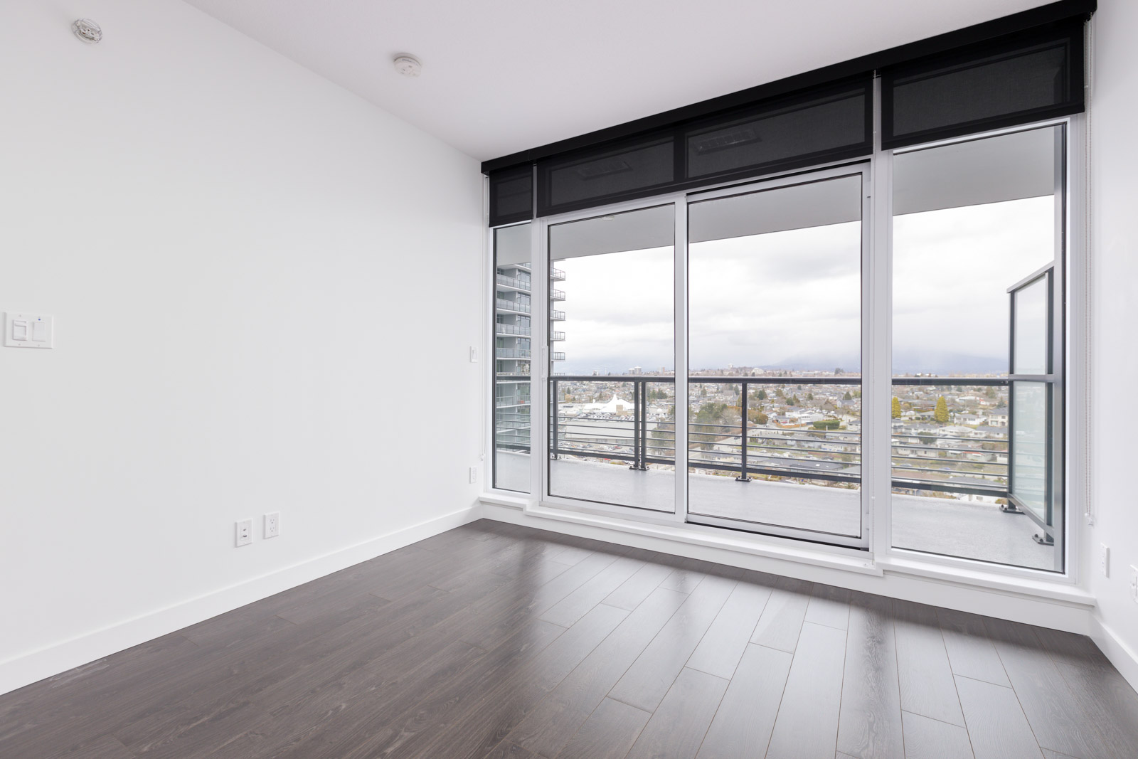 Empty room with dark hardwood flooring, white walls, and a large window leading to a balcony. The window shows a view of a cityscape and overcast sky.