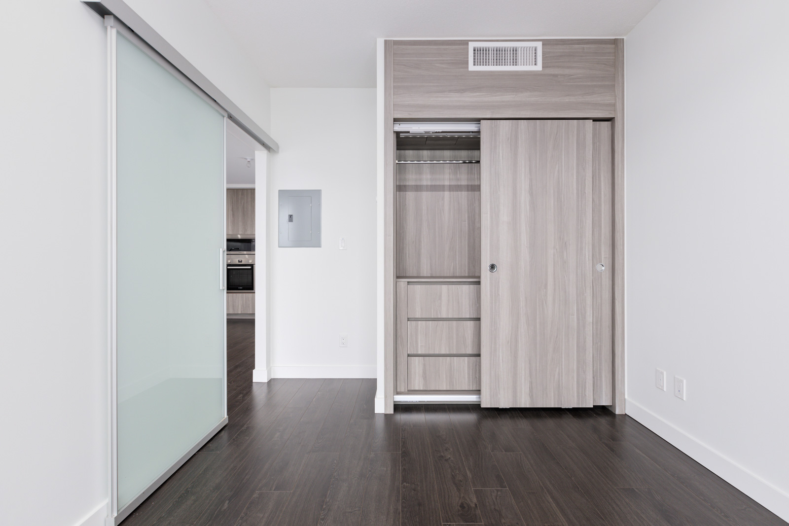 Empty room with dark wood flooring, open closet with shelves and hanging rod, sliding glass door, and visible kitchen area in the background.