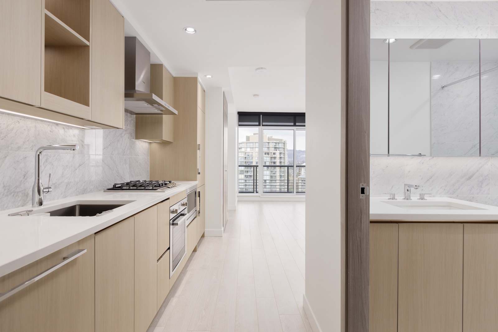 Modern kitchen with light wood cabinets, marble backsplash, and built-in appliances, adjacent to a bathroom and an open living area with large windows and city views.