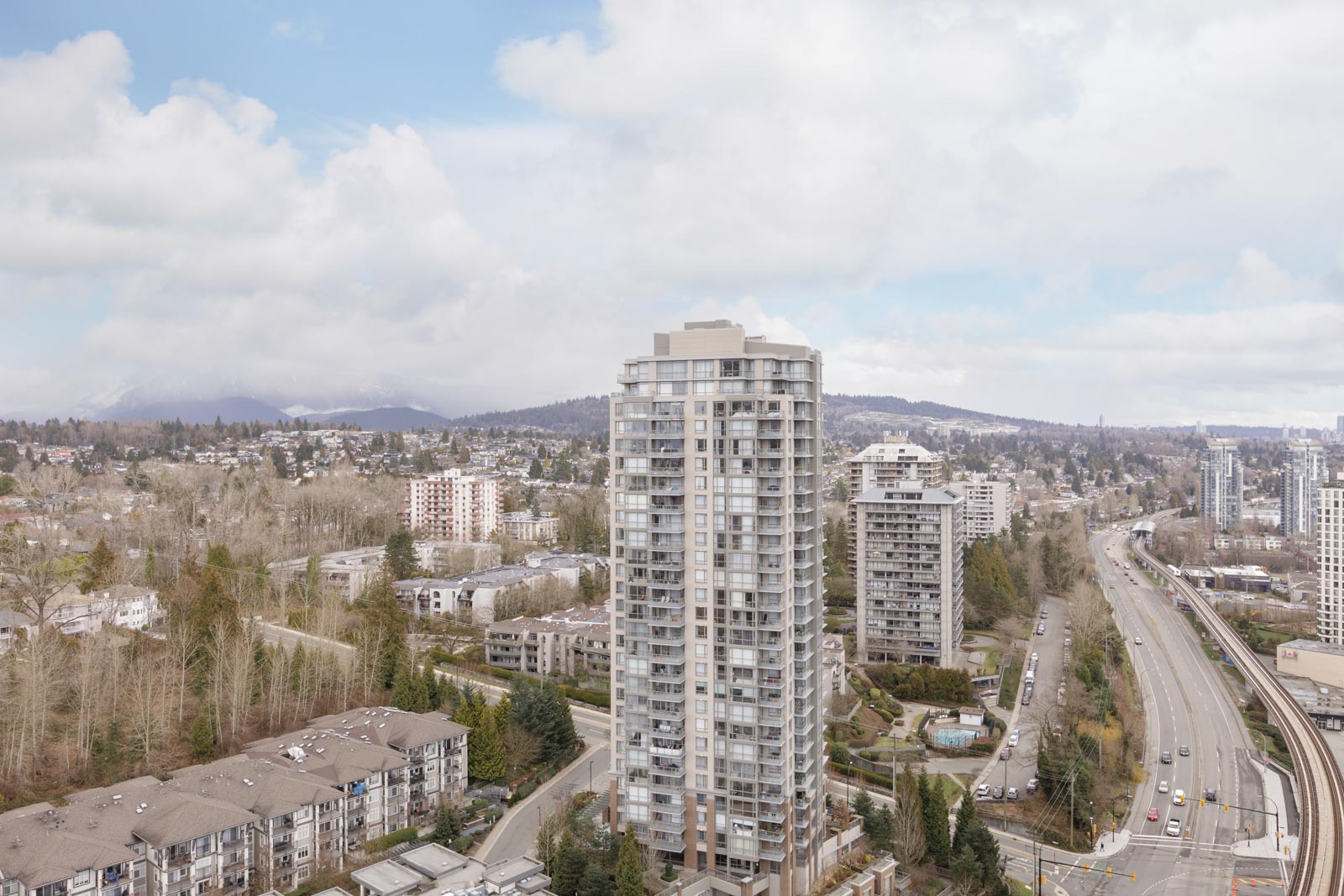 High-rise residential building in the center with roads, smaller buildings, trees, and mountains in the background under a partly cloudy sky.