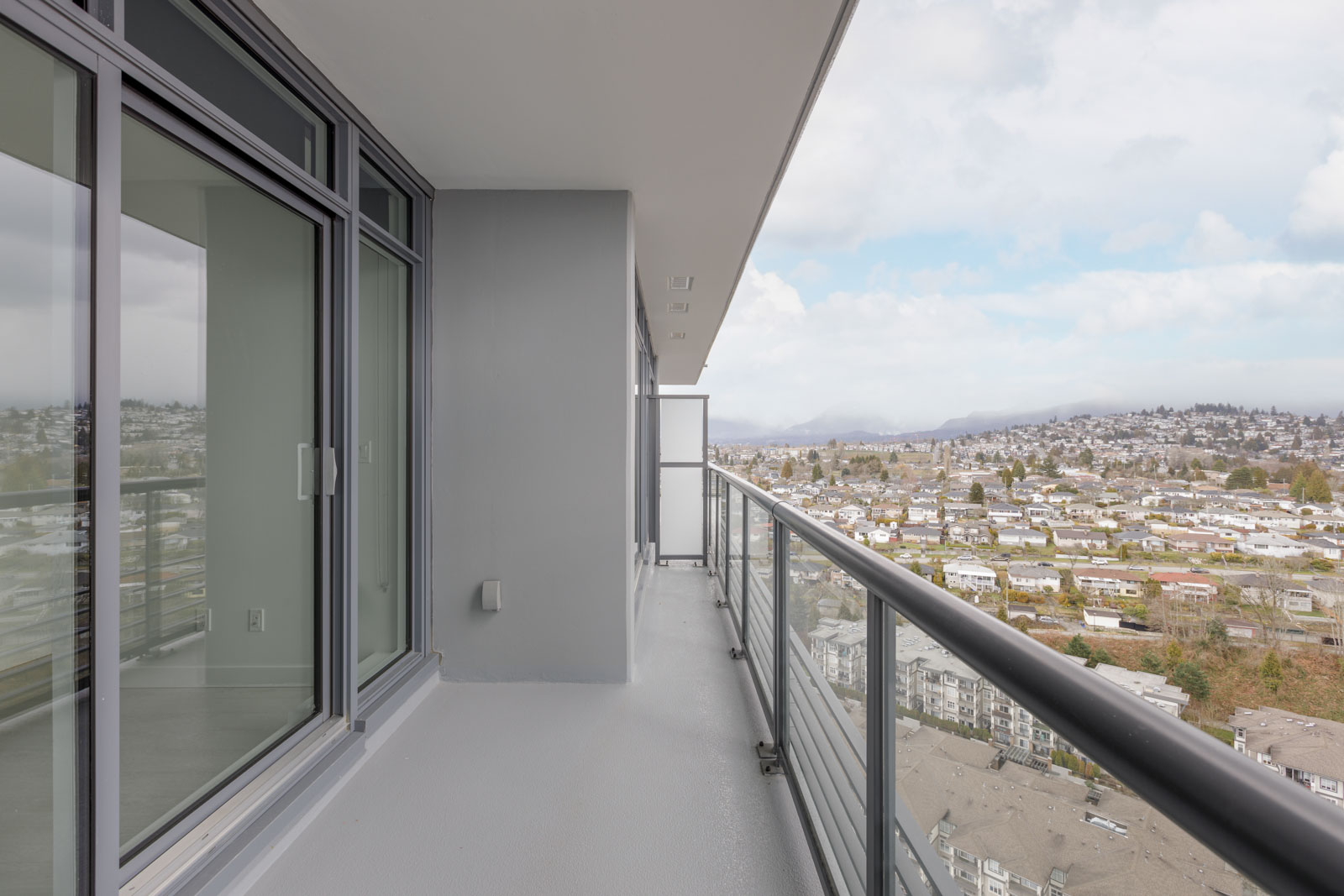 A modern high-rise balcony with glass railing overlooks a cityscape and residential area under a partly cloudy sky.