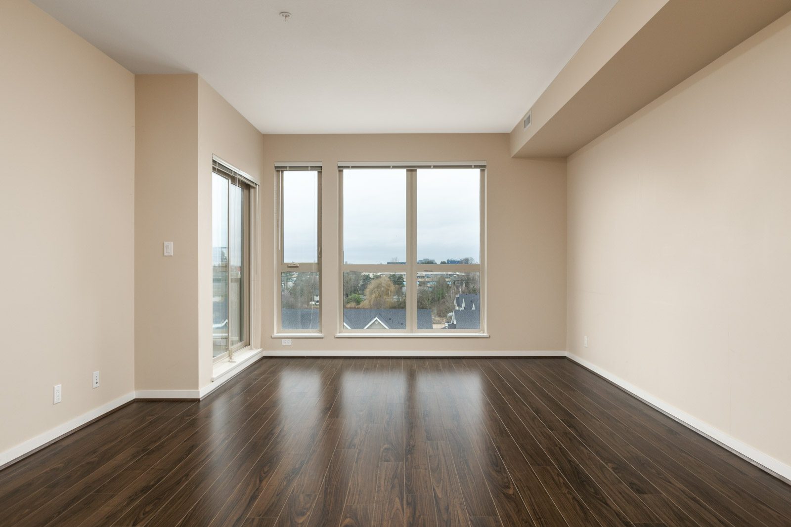 Empty room with large windows, beige walls, and dark wood flooring. It has a view of trees and rooftops outside.