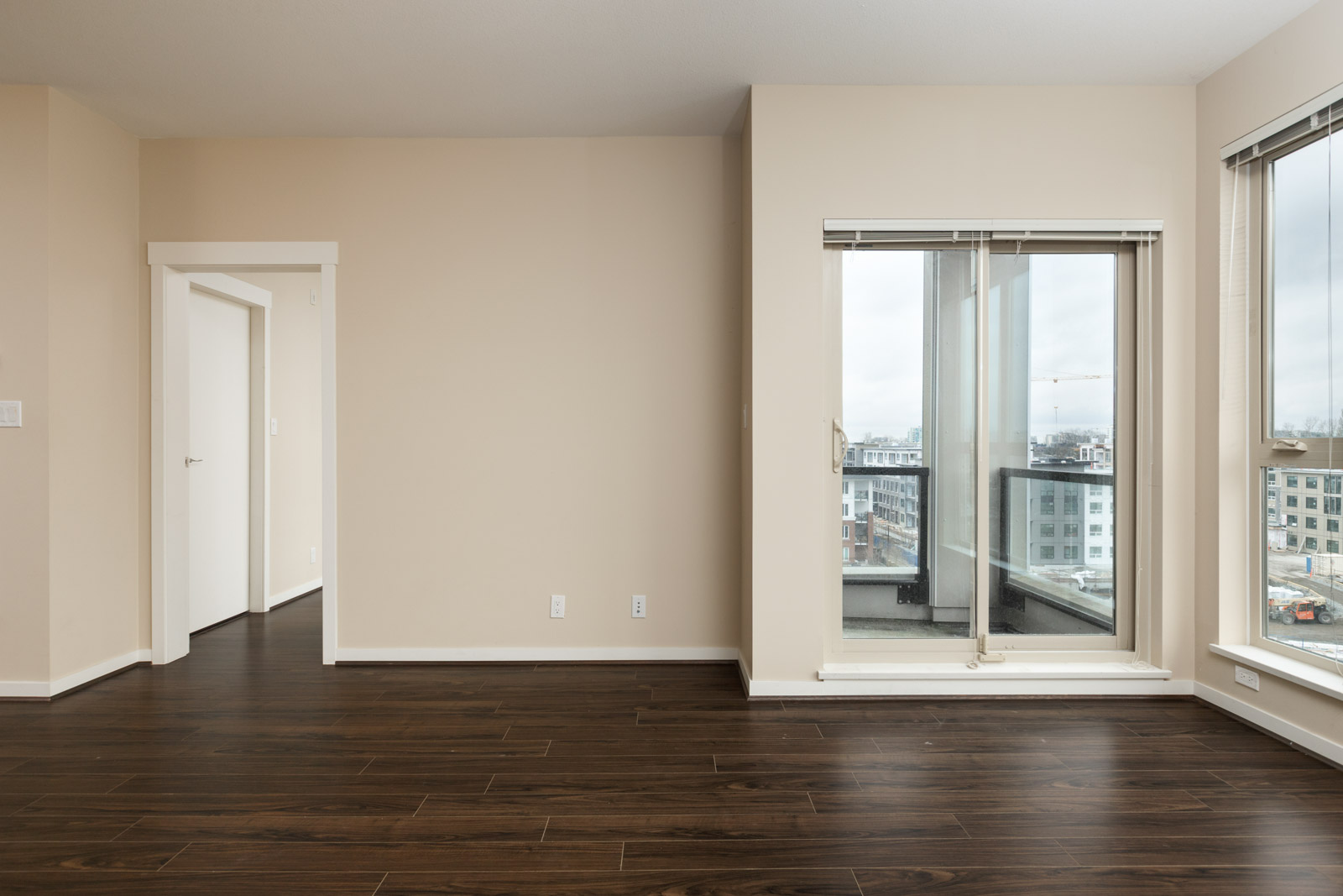 An empty room with beige walls, dark wooden floors, large windows, and a small balcony overlooking a cityscape.