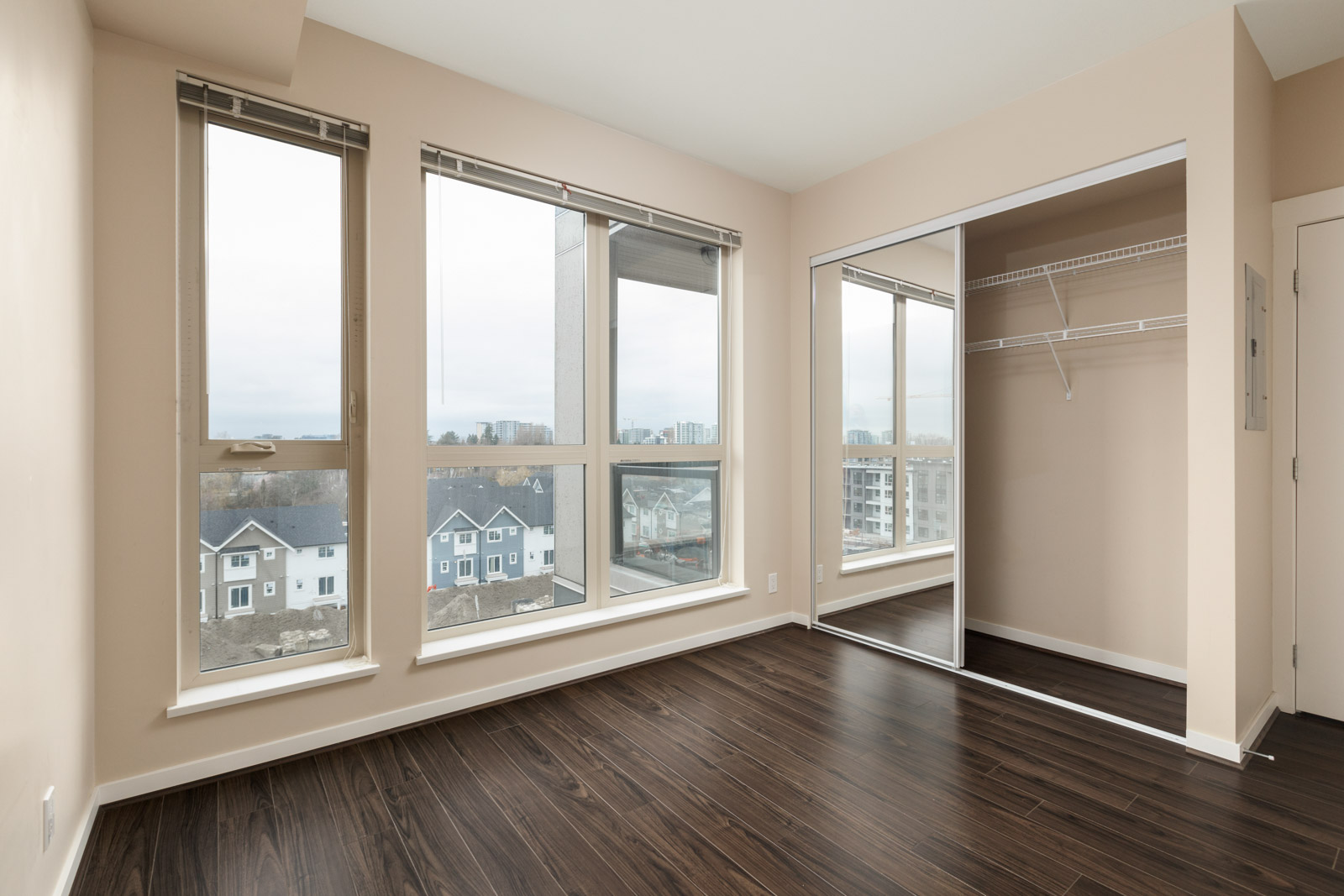 Empty room with large windows and dark wood flooring, overlooking a cityscape. A closet with mirrored sliding doors is on one wall.
