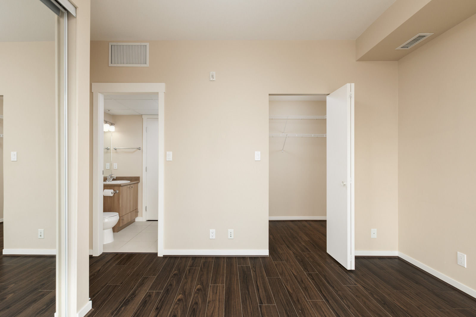 Empty room with dark wood flooring, beige walls, and a white door leading to a closet. An open doorway reveals a bathroom with a sink, mirror, and toilet.