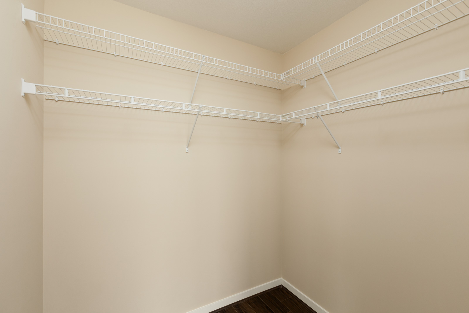 Empty beige walk-in closet with white wire shelving on two walls and a dark wood floor.