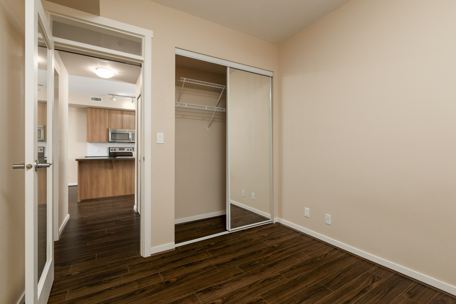 Empty room with wooden floor, a mirrored sliding door closet, and open double glass doors leading to a kitchen area with cabinets and appliances. Walls are beige.