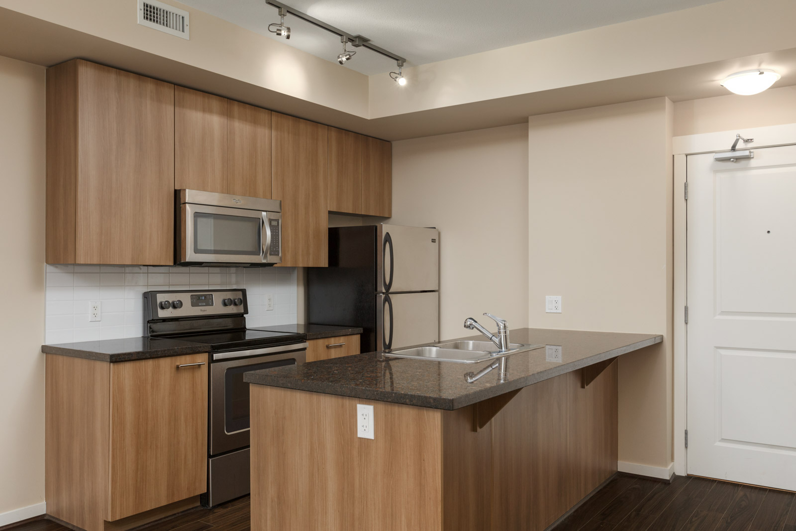 Modern kitchen with wooden cabinets, stainless steel appliances, and a double sink on a black countertop island. Track lighting is on the ceiling, and there's a white door to the right.